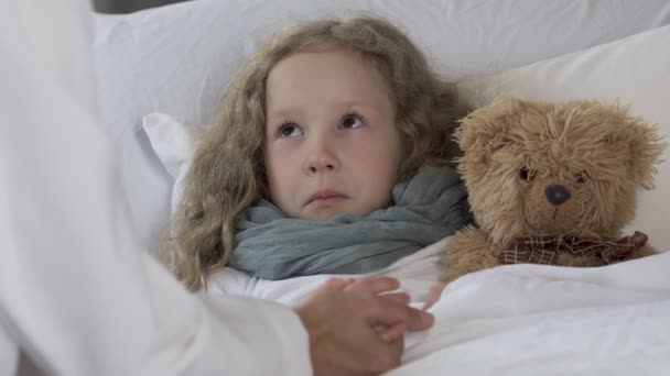 Расстроенная маленькая девочка лежит в постели, имеет высокую температуру и смотрит на врача — стоковое видео