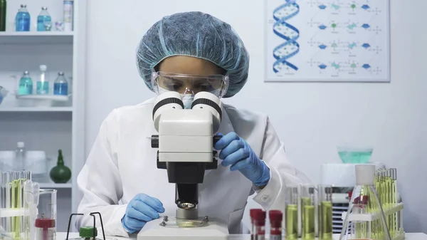 Attentive female lab assistant doing medical research, looking into microscope — Stock Photo, Image