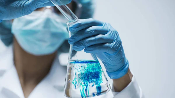 Científico en guantes vertiendo ácido azul en el tubo de ensayo con líquido transparente —  Fotos de Stock