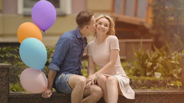 Cara beijando amada namorada na bochecha, segurando balões de ar coloridos na mão — Fotografia de Stock