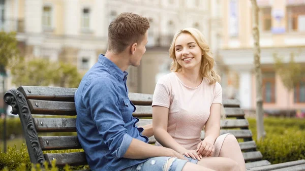 Et ungt par på benken, en fyr som ser på jente og kjærtegner henne, ømhet – stockfoto