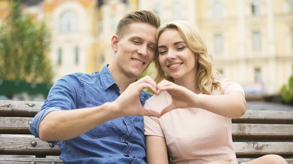 Pareja joven sentada cerca la una de la otra poniendo el corazón en forma de dedo, citas — Foto de Stock