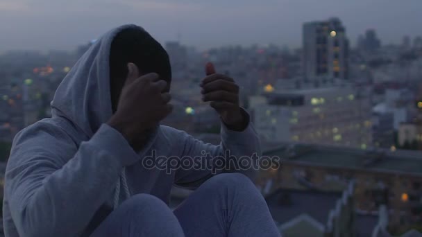 Guy wrapping in sweater sitting on roof edge and peering at night city scenery — Stock Video