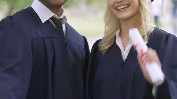 Счастливая пара выпускников в академической одежде, выставляющих свои дипломы — стоковое видео