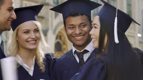 Улыбающиеся выпускники обнимаются, поздравляют с получением дипломов — стоковое видео