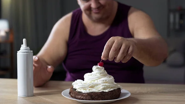 Товстий чоловік кладе вишню на верхівку солодкого торта, прикрашеного збитими вершками — стокове фото