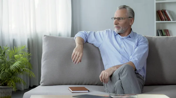 Healthy senior man wearing eyeglasses, sitting on sofa, dreaming about holiday — Stock Photo, Image