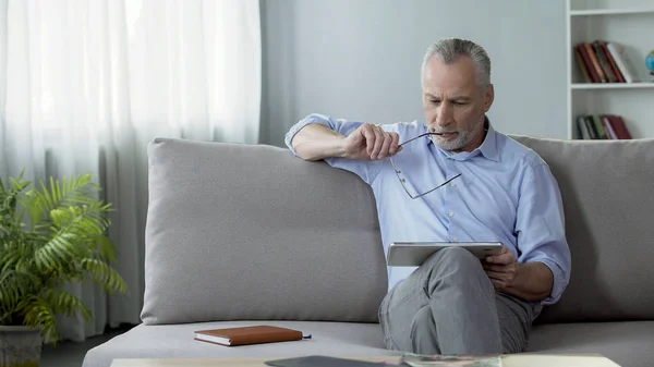 Hombre adulto sentado en el sofá y noticias de lectura en la tableta, las tecnologías modernas — Foto de Stock