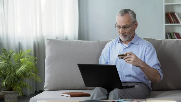 Rentner sitzt auf Sofa und gibt Kartennummer in Laptop ein, Online-Einkauf — Stockfoto