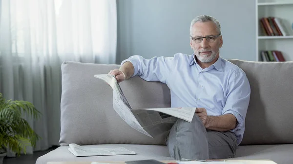 Starší muž sedí na gauči doma, hospodářství noviny a při pohledu do kamery — Stock fotografie