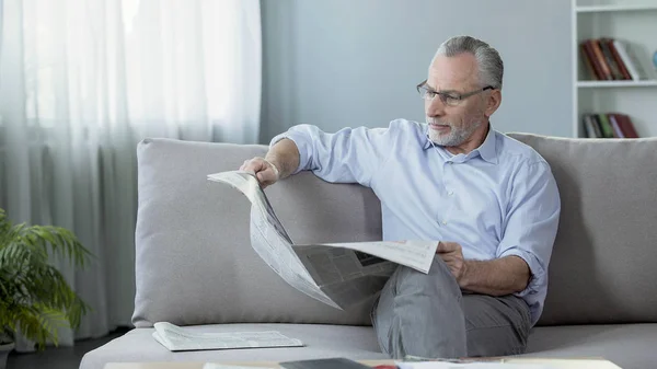 Чоловік у свої 60 років сидить на дивані і читає газету, ранковий ритуал, преса — стокове фото