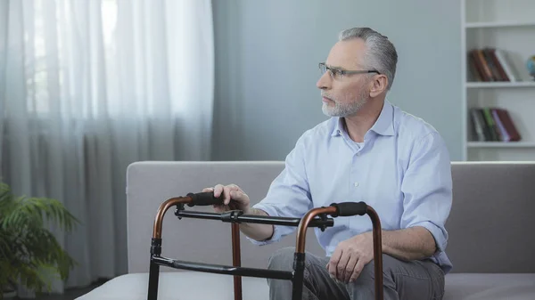 Mann um die 60 sitzt auf Sofa und blickt auf Gehgestell vor sich — Stockfoto