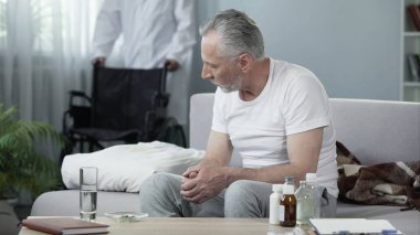Huzurevi, erkek hemşire tekerlekli sandalye getirerek, kanepede oturan özürlü adam