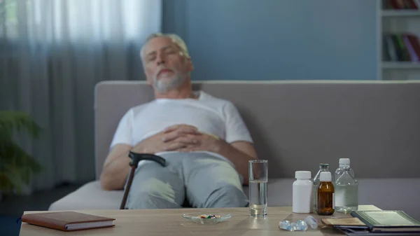 Медикаменти і склянка води, що стоїть на столі, хворий чоловік спить на дивані — стокове фото