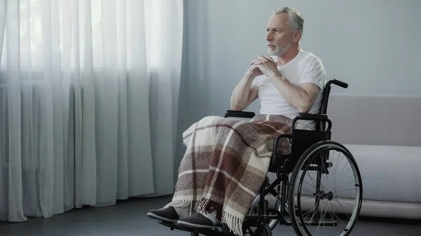 Hombre discapacitado mayor sentado en silla de ruedas y mirando en la ventana, esperando a la familia —  Fotos de Stock