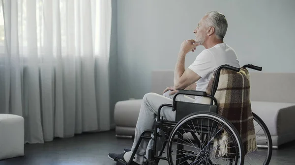 Hombre jubilado reflexivo sentado en silla de ruedas y mirando por la ventana, enfermedad —  Fotos de Stock