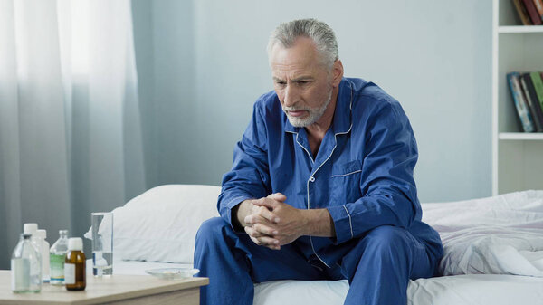 Sad old man sitting in bed and looking at pills, medication and healthcare