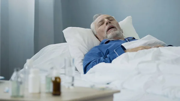 Viejo durmiendo en la cama en el hospital, antibióticos de pie sobre la mesa —  Fotos de Stock