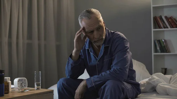 Sénior do sexo masculino sentado na cama e sofrendo de dor de cabeça terrível à noite — Fotografia de Stock