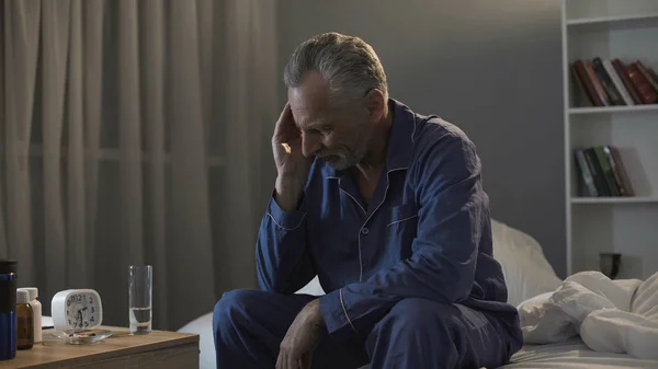 Hombre enfermo retirado en pijama sentado en la cama y sufriendo de dolor de cabeza en la noche —  Fotos de Stock