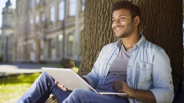 Mixed-race young man with laptop sitting under tree, dating website profile clipart