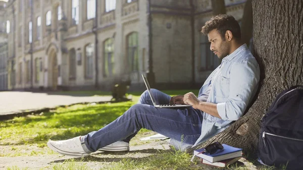 Çok uluslu genç adam ağaç altında laptop hakkında bilgi okuma konsantre