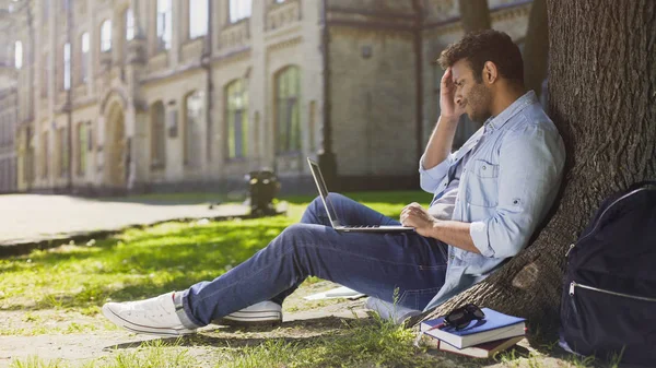 College student siedząc pod drzewem z laptopa szuka zdenerwowany rozczarowujące nowości — Zdjęcie stockowe