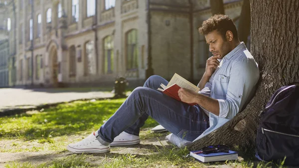 대학생 창과 음모, 몰두와 나무 읽기 책 아래 앉아 — 스톡 사진