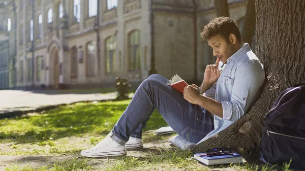 Мультирасовый молодой парень сидит под деревом, читает интересную книгу, книжного червя — стоковое фото