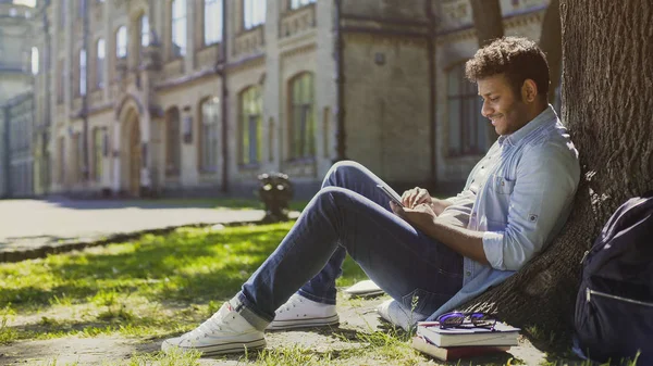 Multiraciale jongeman zittend onder de boom, scrollen scherm van de mobiele telefoon, glimlachend — Stockfoto