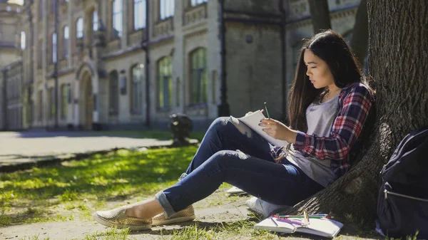 Menina asiática sentada debaixo da árvore, fazendo esboço dos arredores em notebook, artista — Fotografia de Stock