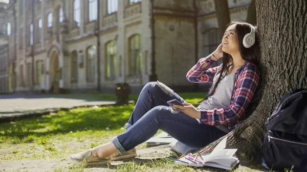 Студентка сидит на траве, прислонившись к дереву, слушает музыку, расслабляется — стоковое фото