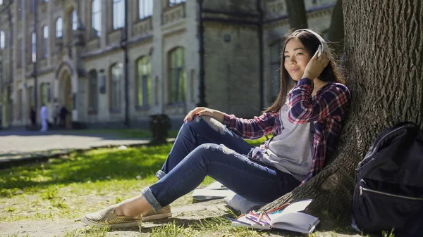 Multinationella ung kvinna i hörlurar som sitter under träd, lyssna på musik — Stockfoto