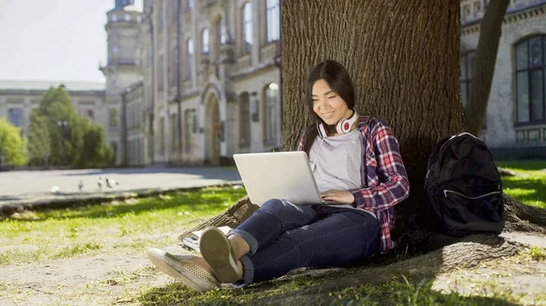 Universitetsstudent sitter under träd i campus, med hjälp av laptop, leende på hennes ansikte — Stockfoto