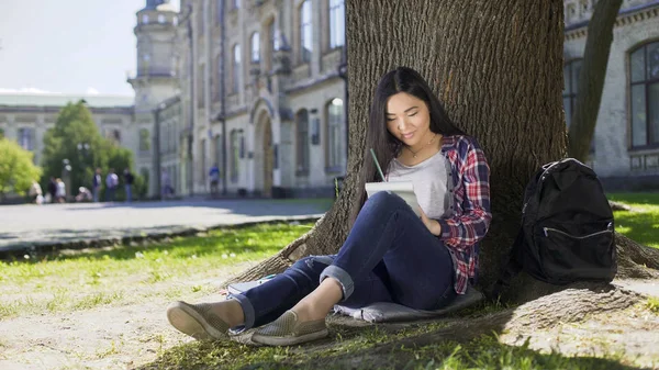 Esej psaní poznámek v poznámkovém bloku, sedící pod stromem, nadnárodní mladá žena — Stock fotografie