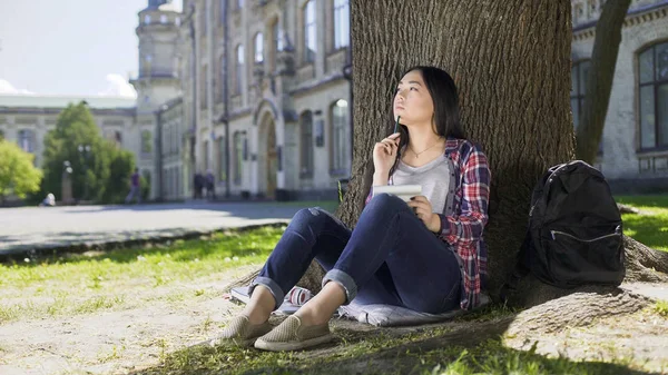Výměnný student sedí pod stromem, drží notebook, myslet na projektu — Stock fotografie