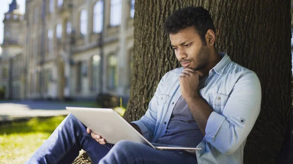 Junger Mann mit gemischter Rasse sitzt unter Baum und schaut auf Laptop, wichtiger Brief — Stockfoto