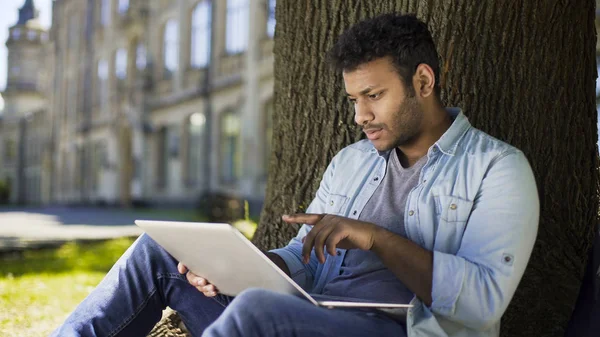 Multinacional joven sentado bajo el árbol, mirando portátil lectura oferta de trabajo —  Fotos de Stock