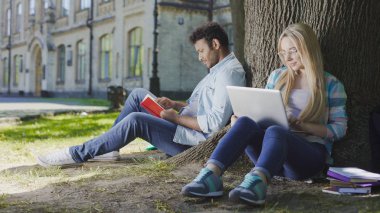 Genç erkek erkek dizüstü bilgisayar, öğrenci yaşamı ile yakınındaki kitapla ağacın altında oturup