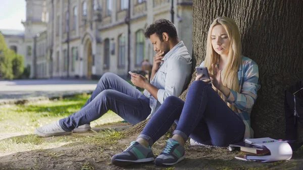 Női, férfi idegenek fa alatt ült, használja a mobilját, látszó lány aggódik — Stock Fotó