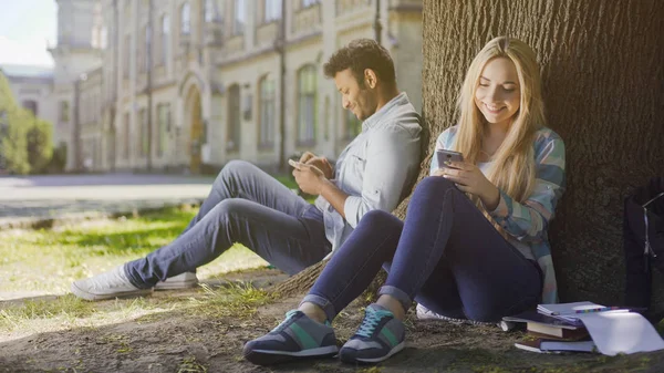 Jovens sentados debaixo da árvore, usando celular, sorrindo para si mesmos, deleitam-se — Fotografia de Stock