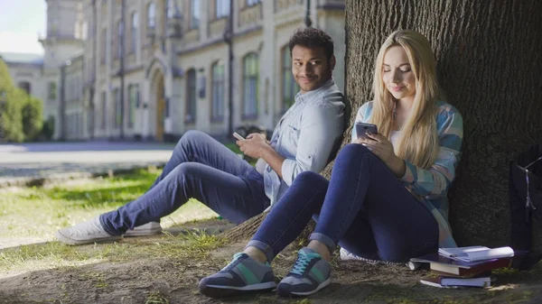 Mies, jolla on kännykkä ja joka istuu puun alla ja katselee tyttöä puhelimen, kiintymyksen — kuvapankkivalokuva
