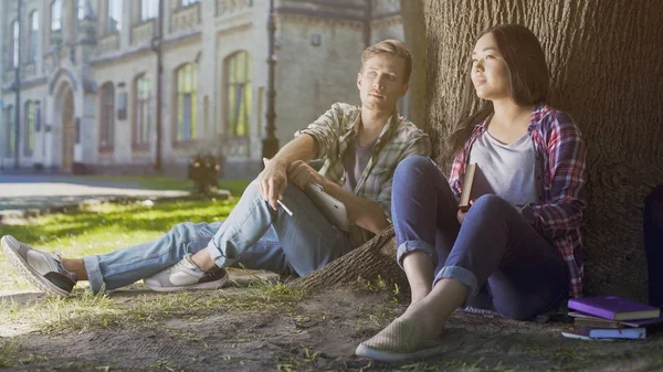 Багаторасові студентки чоловічої та жіночої статі сидять під деревом, сподіваючись на майбутнє — стокове фото