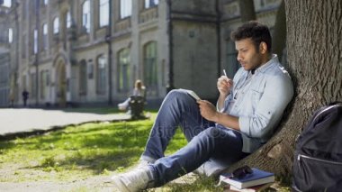 Çok ırklı erkek ağaç altında çimlerin üzerine oturan defter, yaratıcı bir fikir yazma