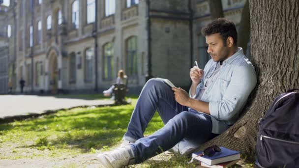 Multiracial male sitting on grass under tree, writing in notebook, creative idea — Stock Video