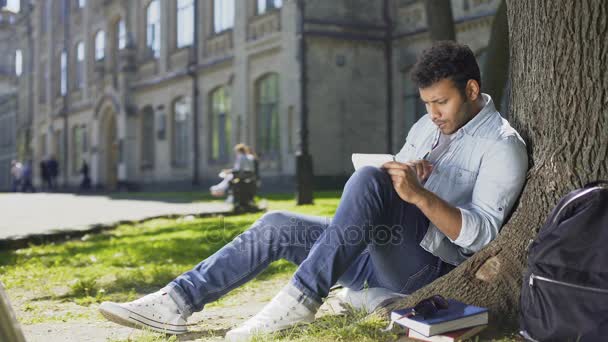 Φοιτητής πανεπιστημίου που κάθεται κάτω από το δέντρο στην πανεπιστημιούπολη, λύνοντας την εργασία στο σημειωματάριο, έξυπνη — Αρχείο Βίντεο