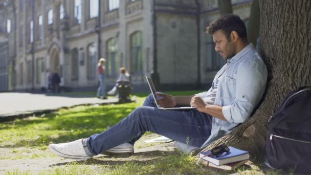 Estudiante universitario sentado bajo el árbol, usando el ordenador portátil mirando preocupados, noticias perturbadoras — Vídeos de Stock