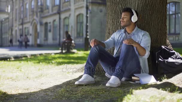 Молодой человек кладет наушники, слушает музыку сидя под деревом, восхищение — стоковое видео
