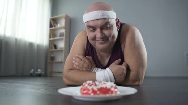 Homem corpulento engraçado deitado no chão e olhando para donut com amor, força de vontade — Vídeo de Stock