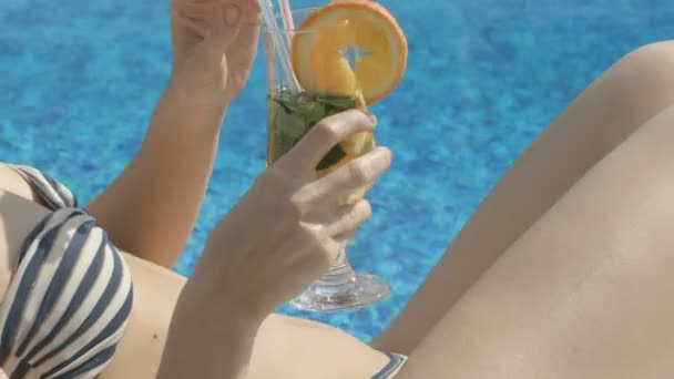 Тонкая леди пьет освежающий коктейль, принимая солнечную ванну у просторного бассейна — стоковое видео